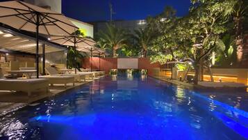 Une piscine nocturne, dotée de chaises longues et de parasols, entourée de palmiers et bordée d’une architecture moderne.