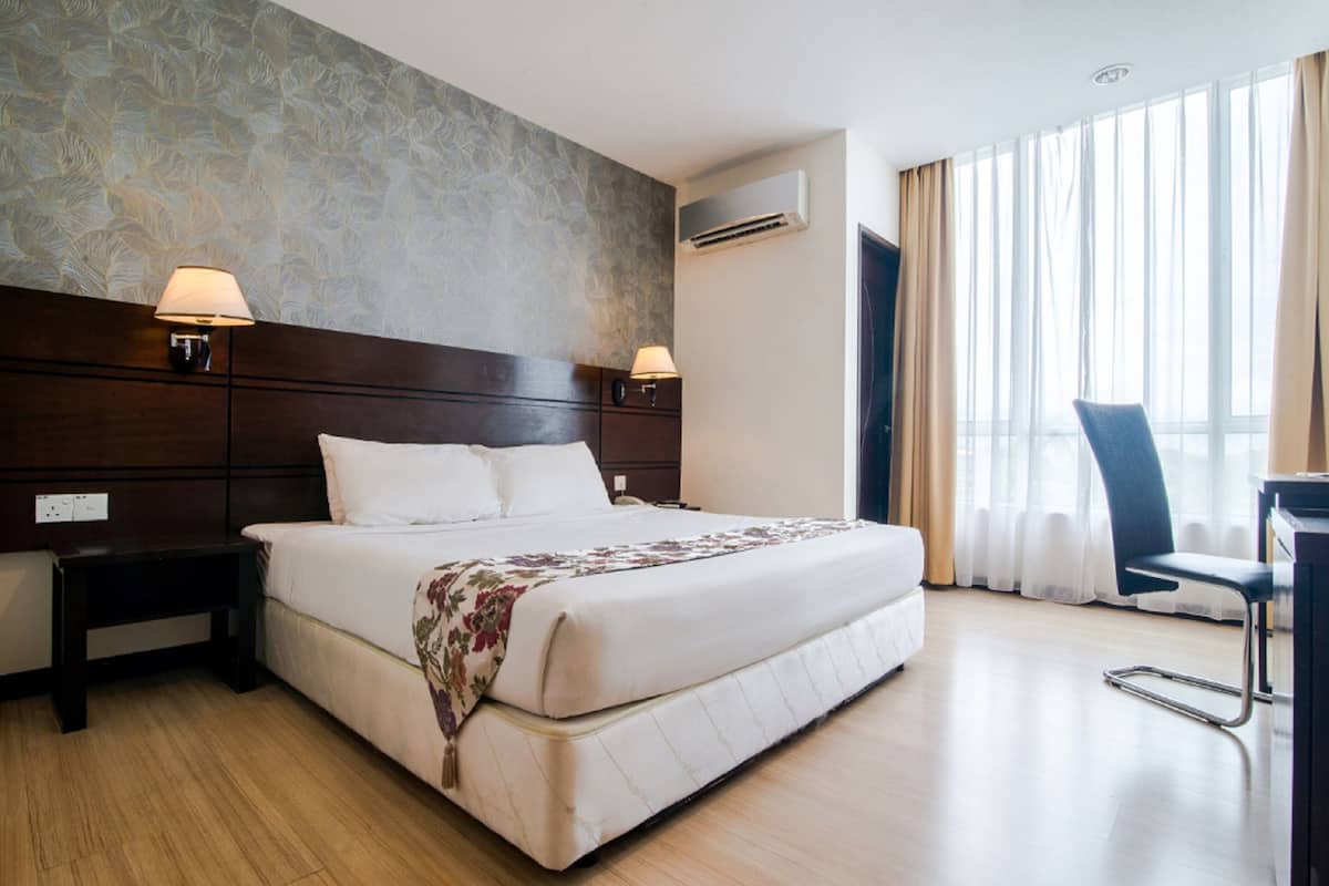 A hotel room with a large bed, two bedside lamps, a wooden headboard, an air conditioning unit, a desk with a chair, and a large window with curtains.
