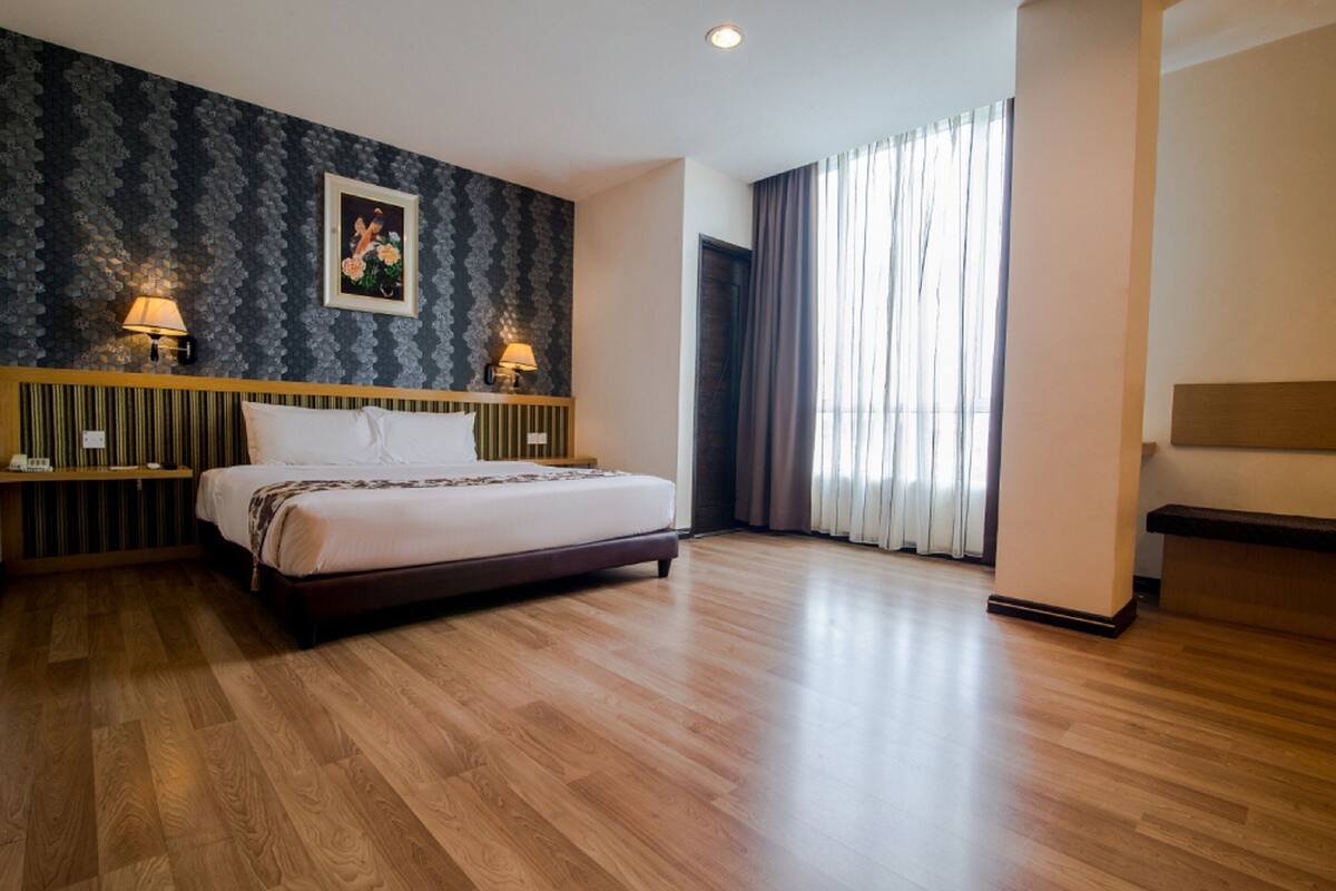 A hotel room with a large bed, two bedside lamps, a framed picture on the wall, and a view of the outdoors through a window with curtains.