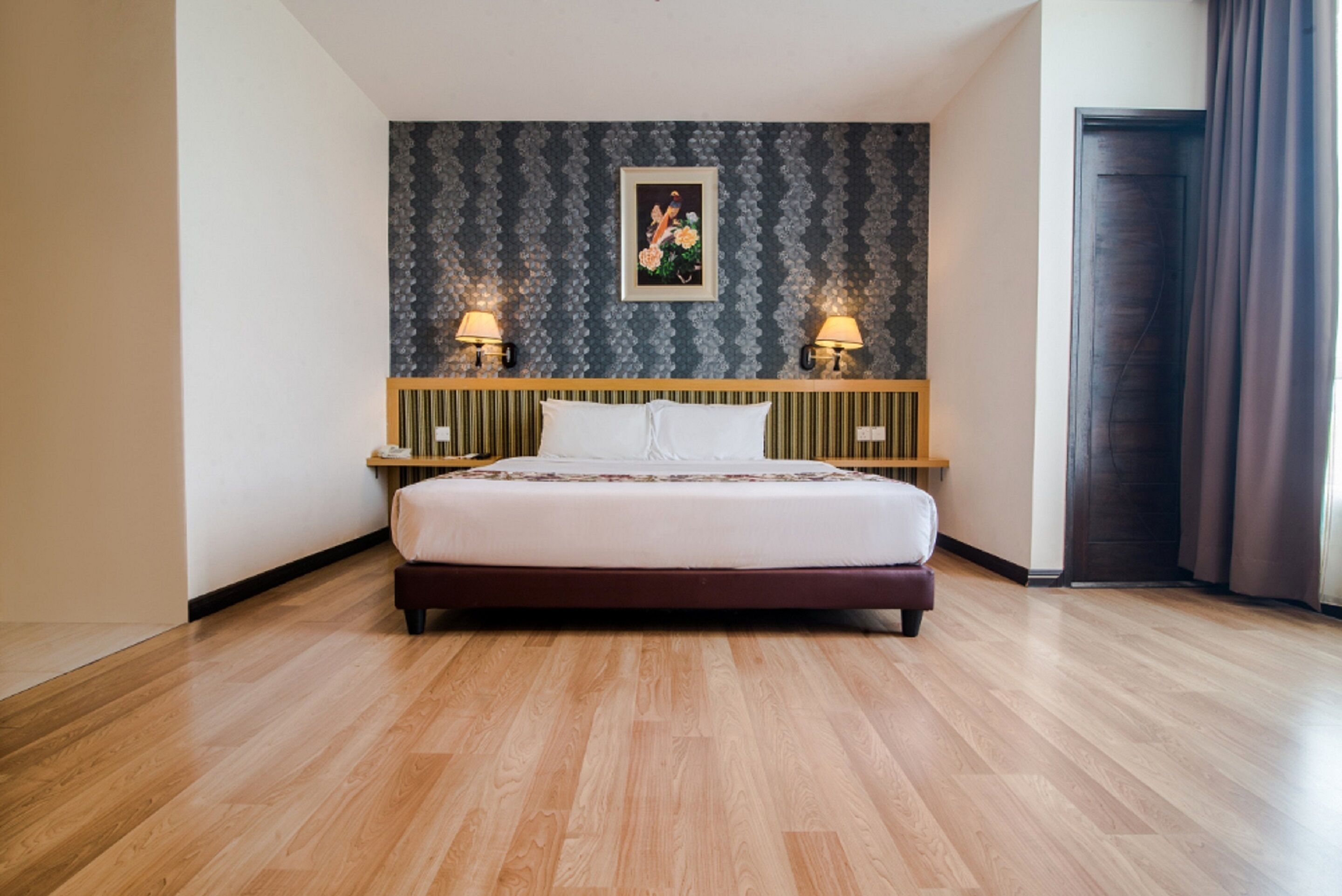 A hotel room with a large bed, wooden flooring, a patterned wall, and two bedside lamps.