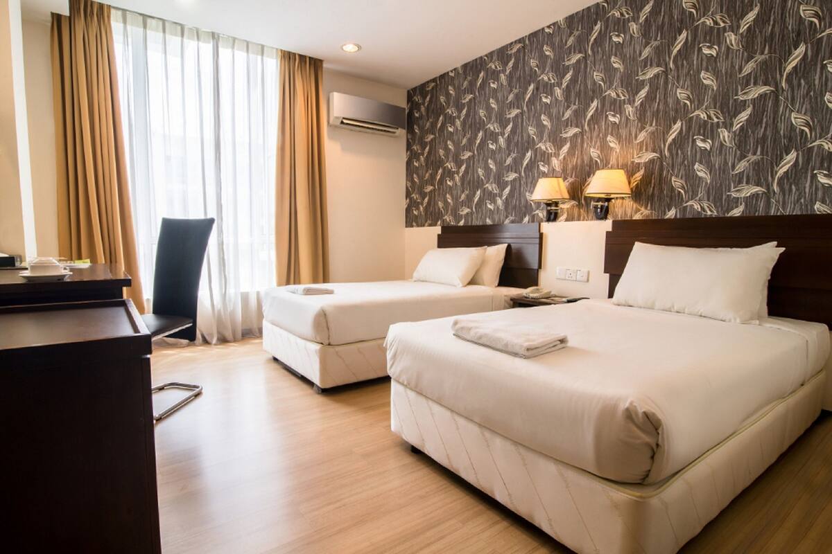 A hotel room with two beds, a desk, and a chair. There is a window with curtains, an air conditioner, and a wall with a leaf pattern.