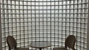 A room with a curved wall of transparent glass blocks, a round table, and two chairs.