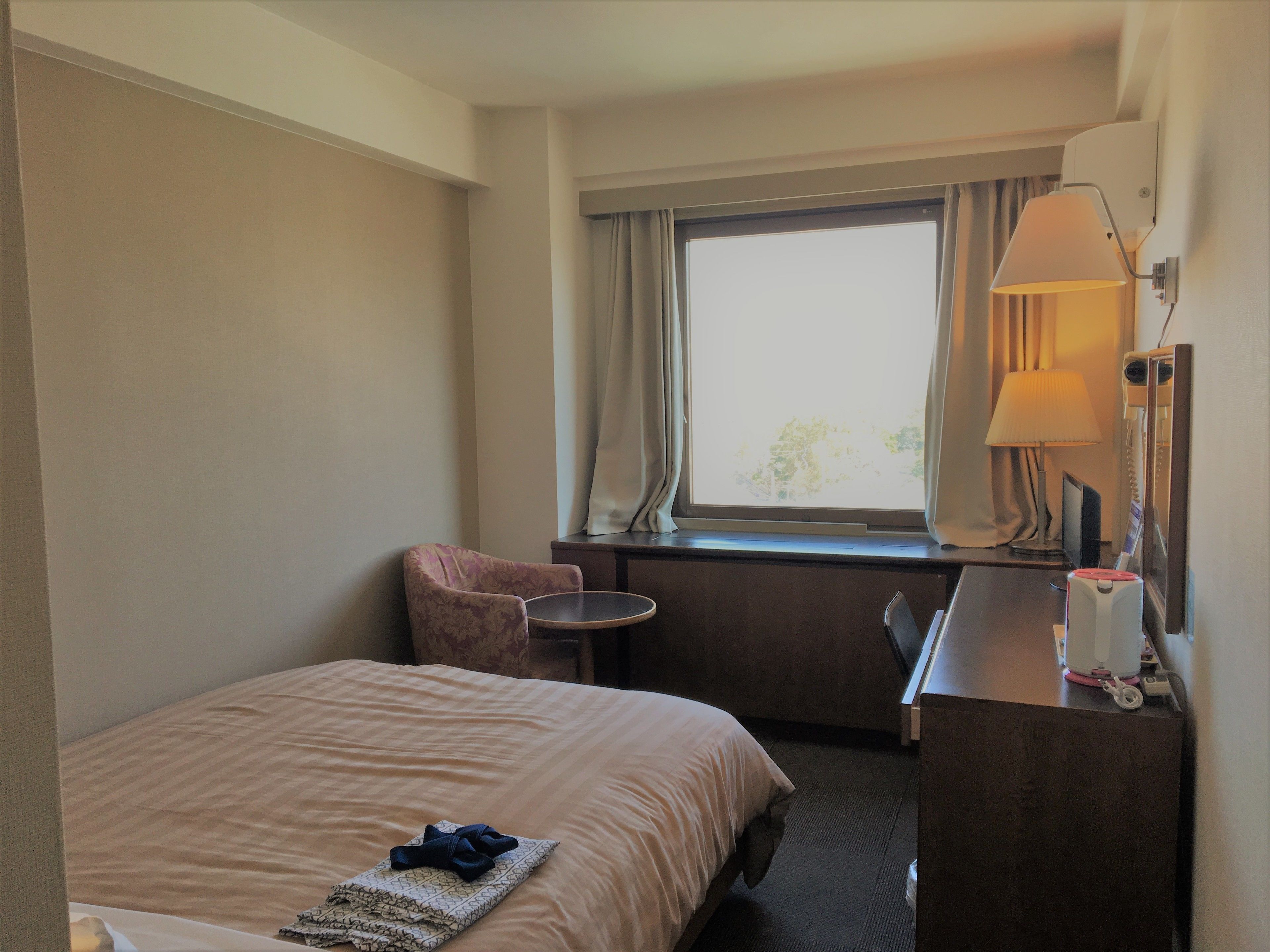 A hotel room with a bed, a chair, a desk, a lamp, and a window with curtains.