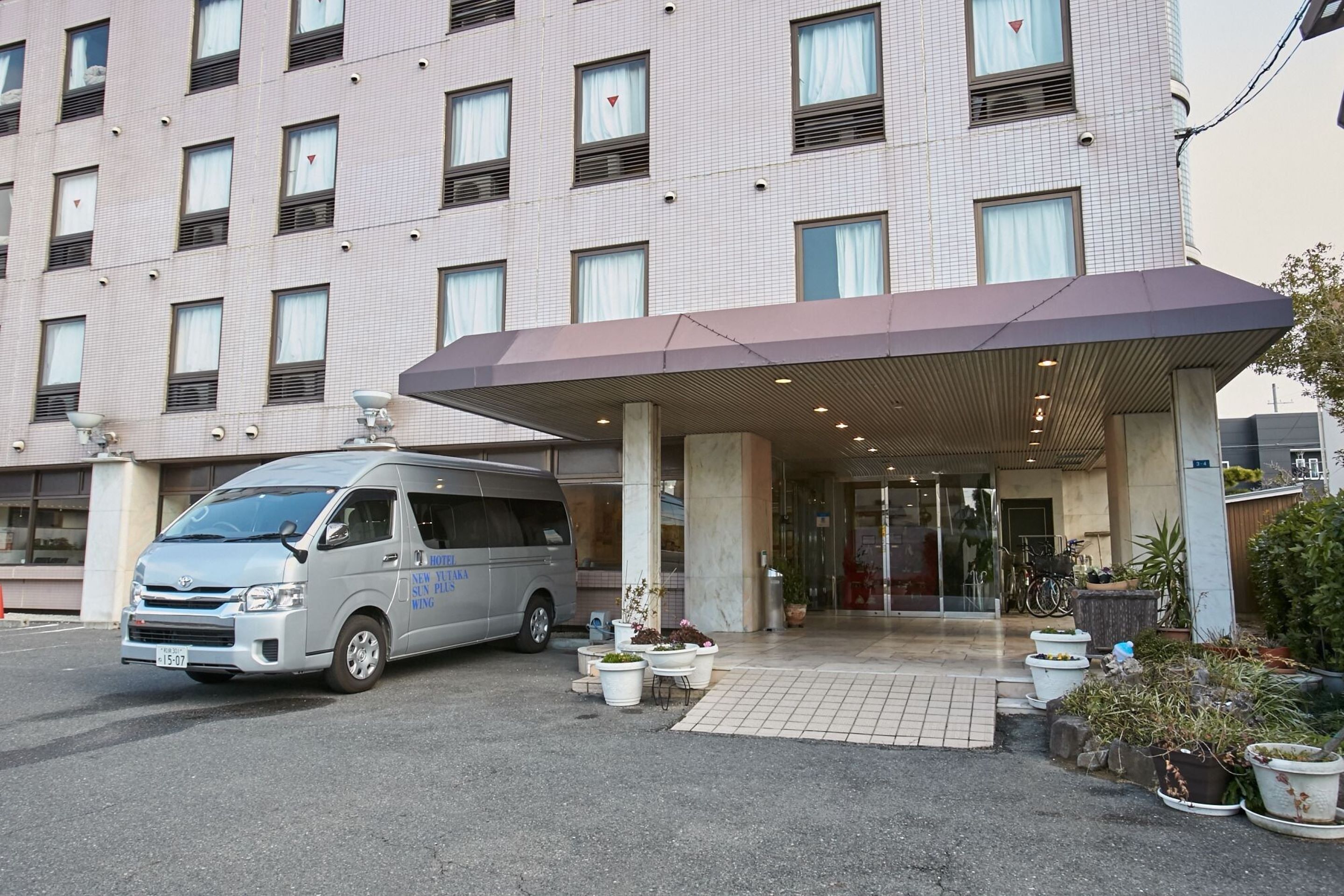 A hotel entrance with a van parked in front, potted plants, and a covered entrance area.