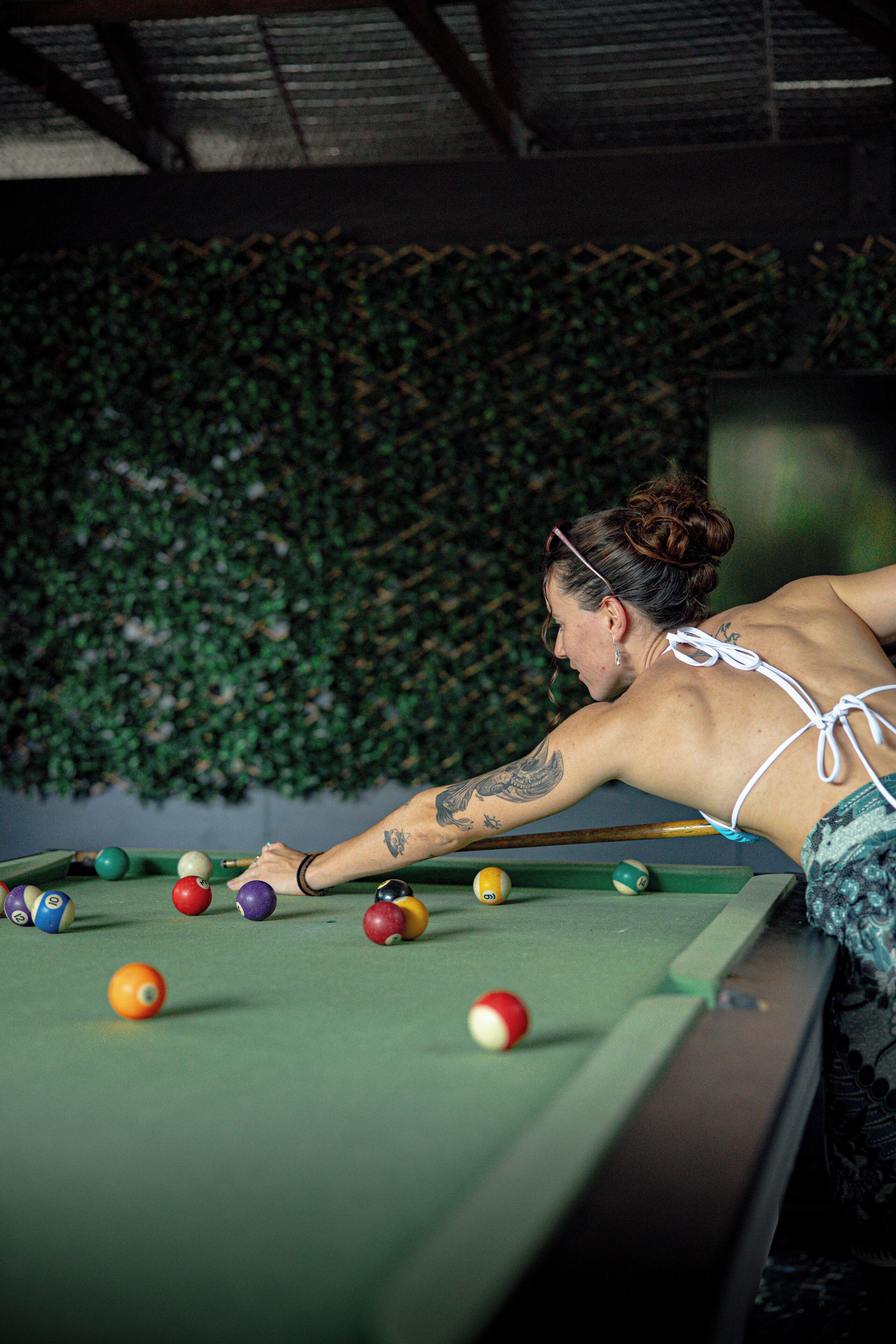 A person playing pool in a room with a green pool table and a wall covered with green foliage.