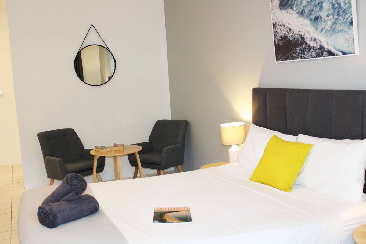 Quarto de hotel com cama, duas poltronas, uma mesinha, espelho e um quadro pendurado na parede.
