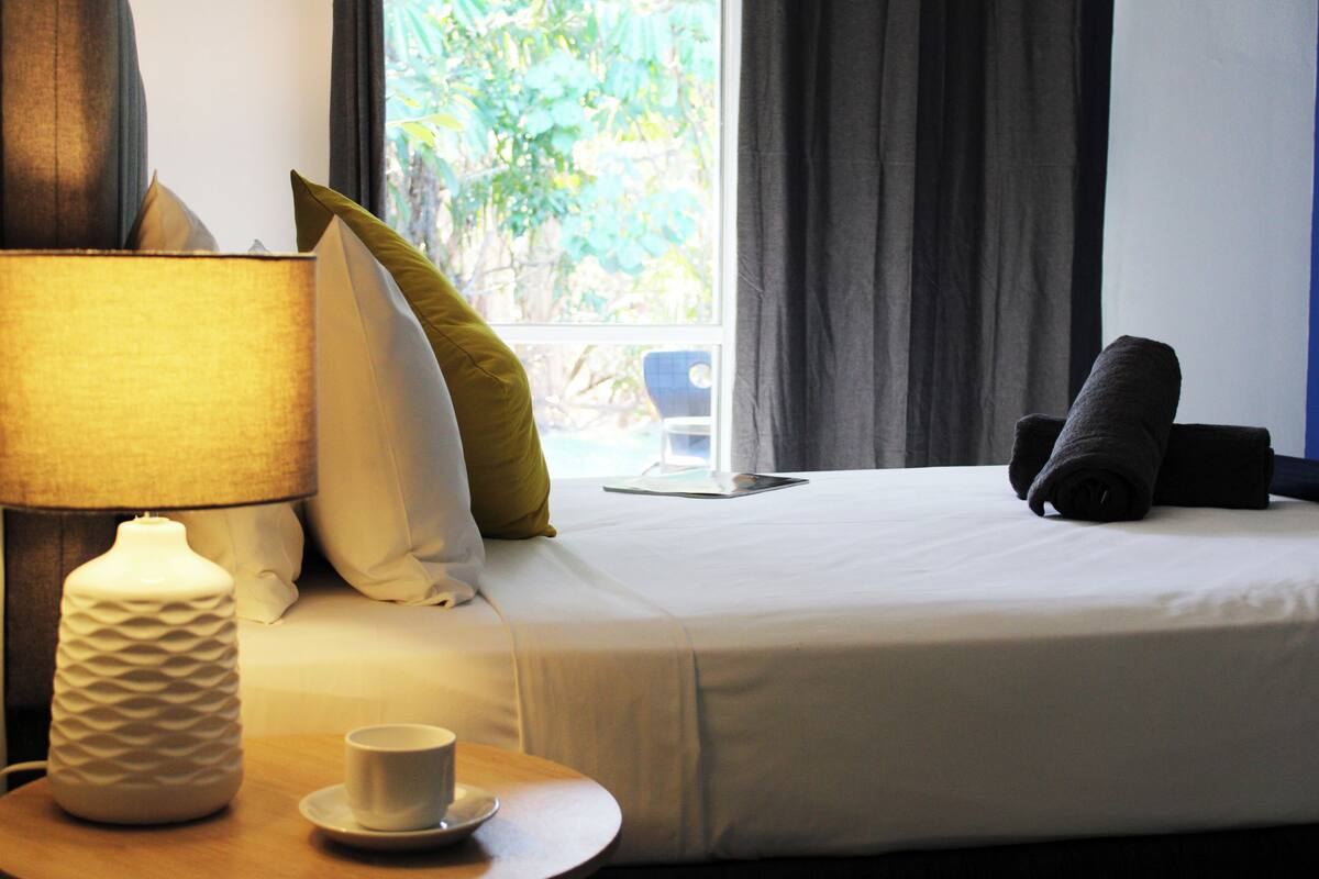 Quarto de hotel com uma cama, mesa de cabeceira com abajur, uma xícara e vista para o verde lá fora.