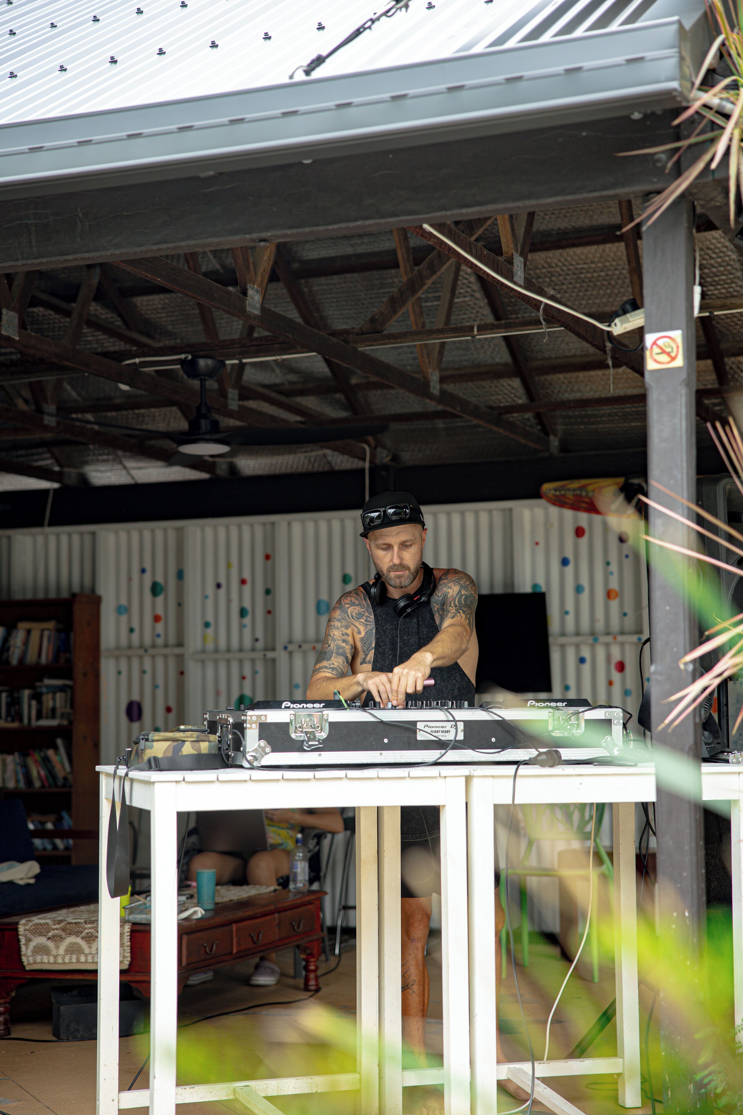 Eine Person sitzt an einem DJ-Pult mit elektronischer Ausrüstung, in einem Raum mit Wellblechdach und einem Bücherregal im Hintergrund.