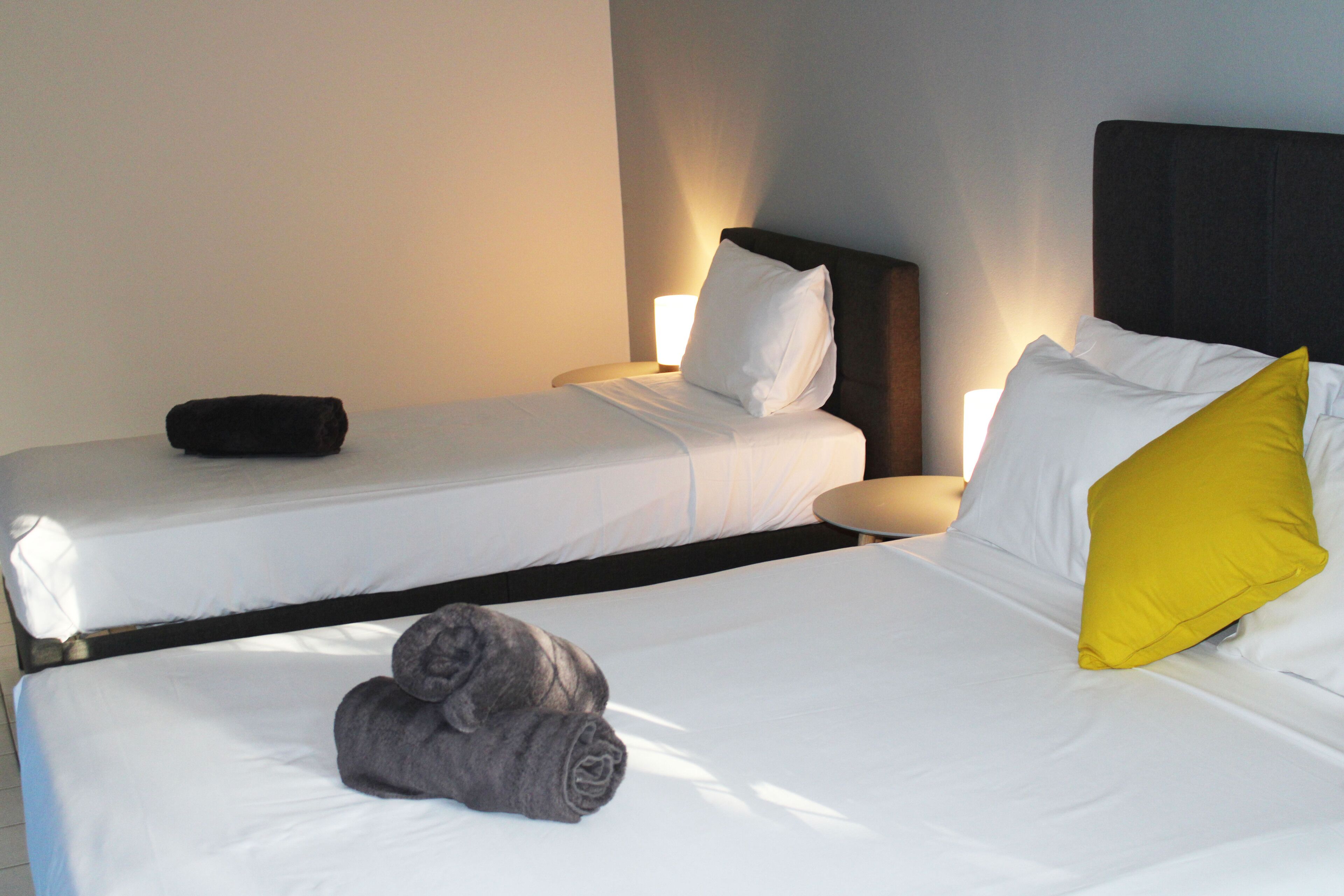 A hotel room with two beds, each with white linens and pillows, one with a yellow pillow. There is a black pillow on the left bed, and a rolled-up gray towel on the floor.