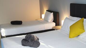 A hotel room with two beds, each with white linens and pillows, one with a yellow pillow. There is a black pillow on the left bed, and a rolled-up gray towel on the floor.