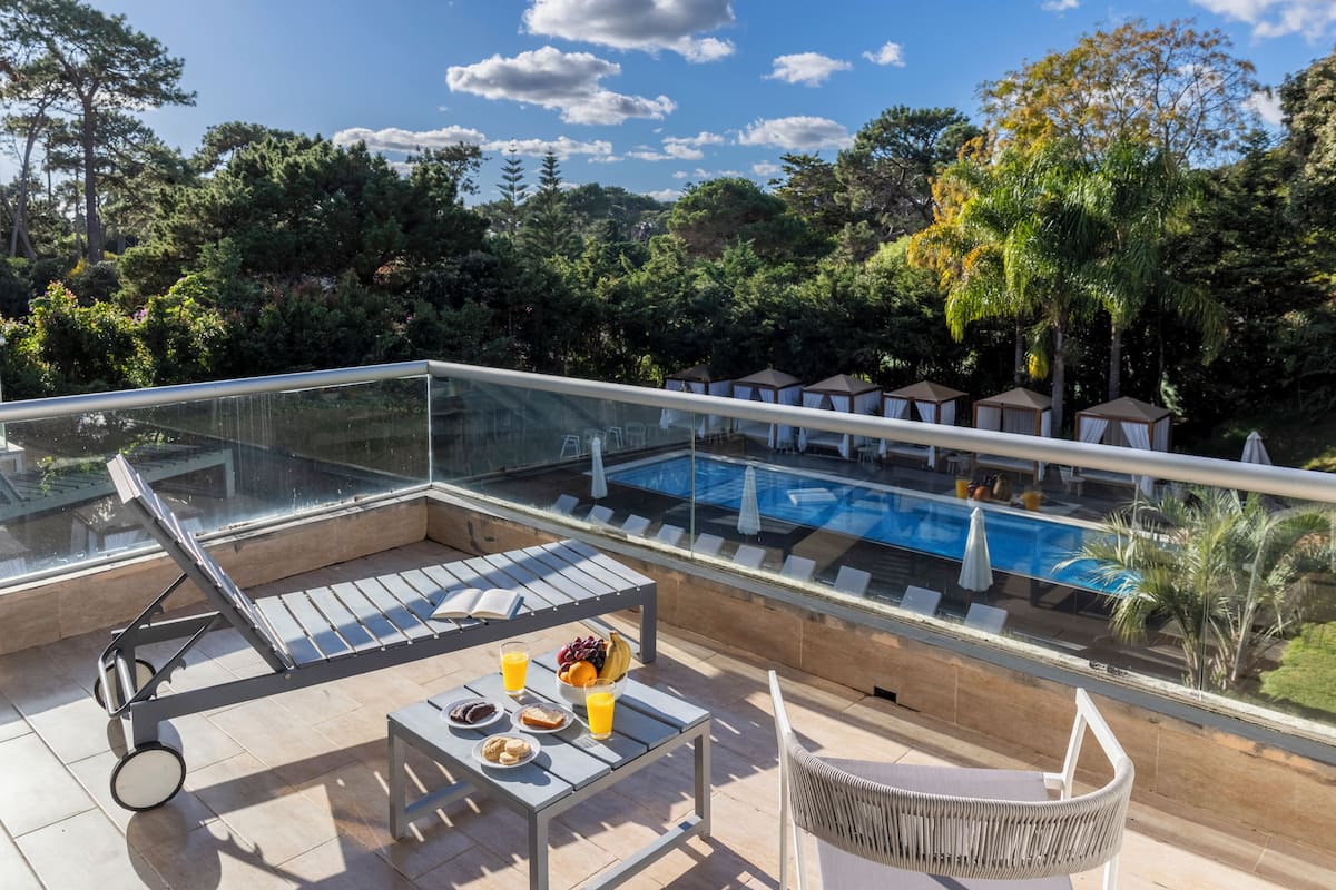 Un espace piscine aménagé sur le toit, avec des chaises longues, une table garnie de fruits et de boissons, et une balustrade en verre offrant une vue sur une piscine et une végétation luxuriante.