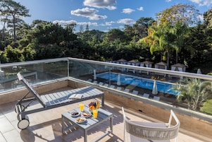 Zona de piscina en la azotea con sillones de descanso, una mesa con fruta y bebidas, y una barandilla de cristal con vistas a una piscina y exuberante vegetación.