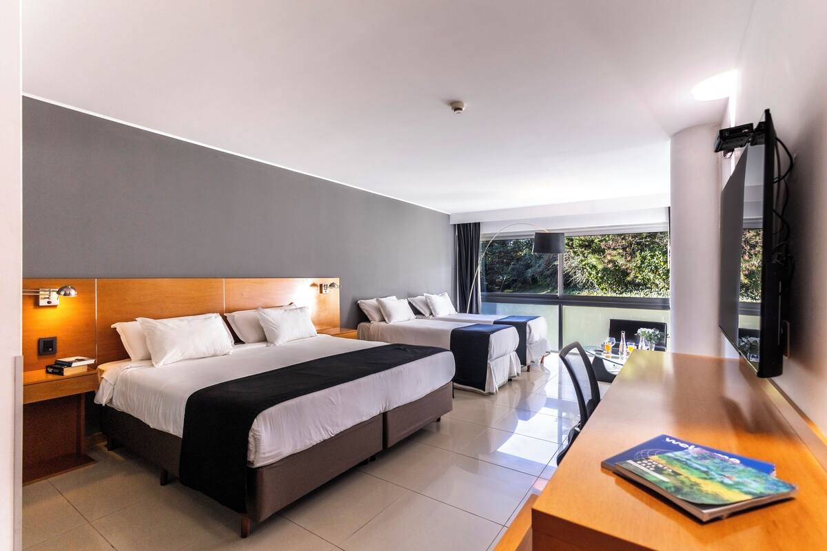 Une chambre d’hôtel moderne dotée de deux lits, d’un bureau en bois, d’une télévision à écran plat et d’une grande fenêtre donnant sur un paysage verdoyant.