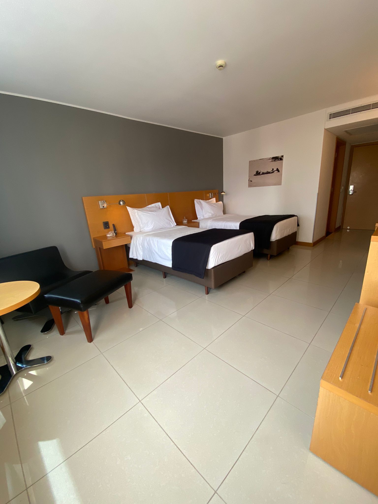 Quarto de hotel com duas camas, uma escrivaninha, uma cadeira e uma mesinha.