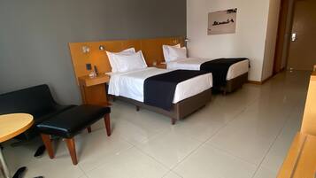 Quarto de hotel com duas camas, uma escrivaninha, uma cadeira e uma mesinha.