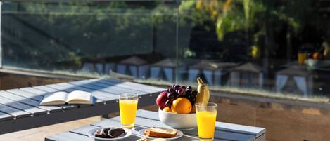 Una mesa con alimentos para desayuno, como pan, galletas y un bol de fruta y jugo, en una terraza al aire libre con vistas a árboles y un edificio al fondo.