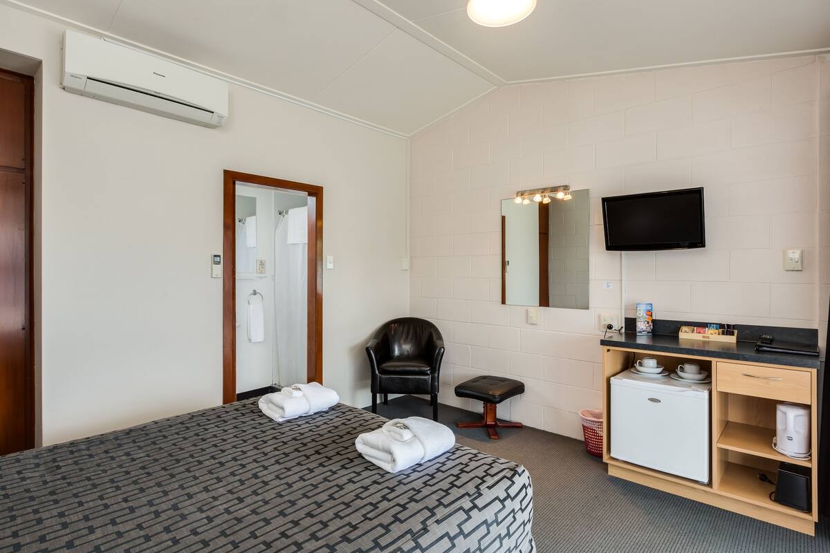 A hotel room with a bed, a chair, a small kitchenette, a TV, and an air conditioning unit.