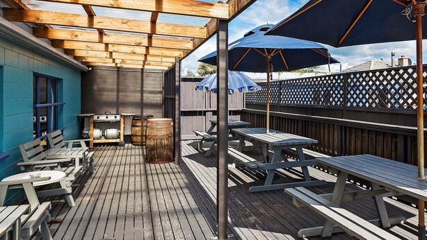 A wooden deck with picnic tables and umbrellas, a barbecue grill, and a blue building in the background. - Ocean Beach Hotel (Dunedin)