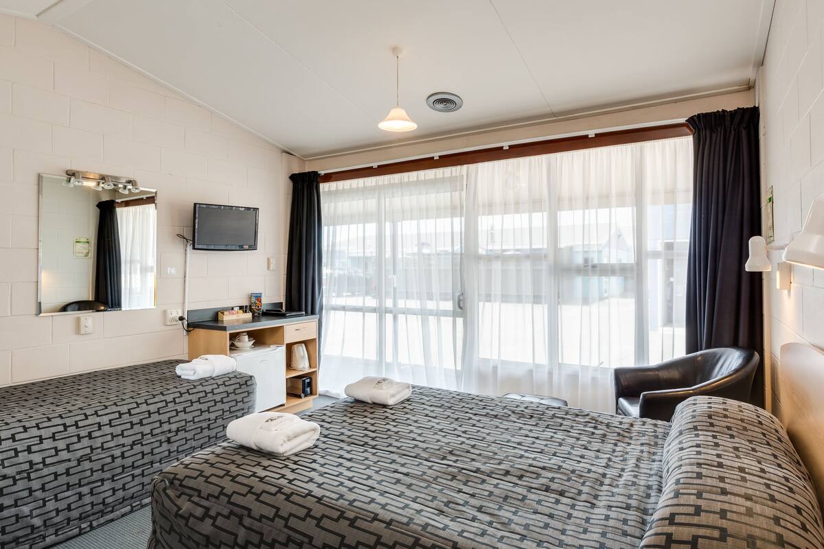 A hotel room with two beds, a desk, a chair, a TV, and a large window with curtains.