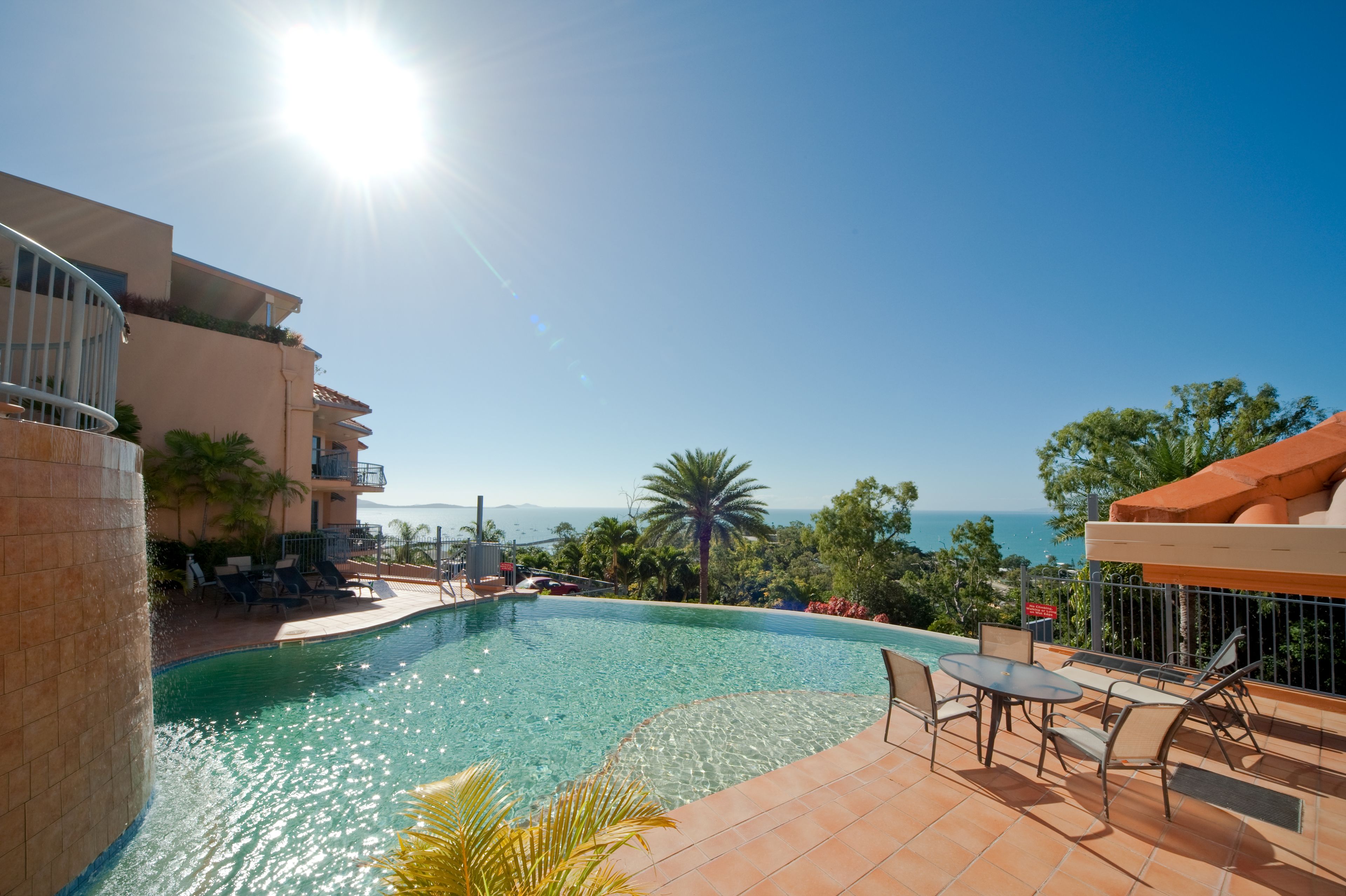 A swimming pool with a view of the ocean, surrounded by palm trees and a building with balconies.