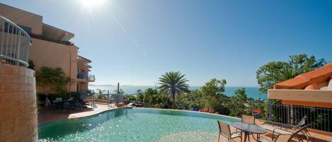 A swimming pool with a view of the ocean, surrounded by palm trees and a building with balconies.