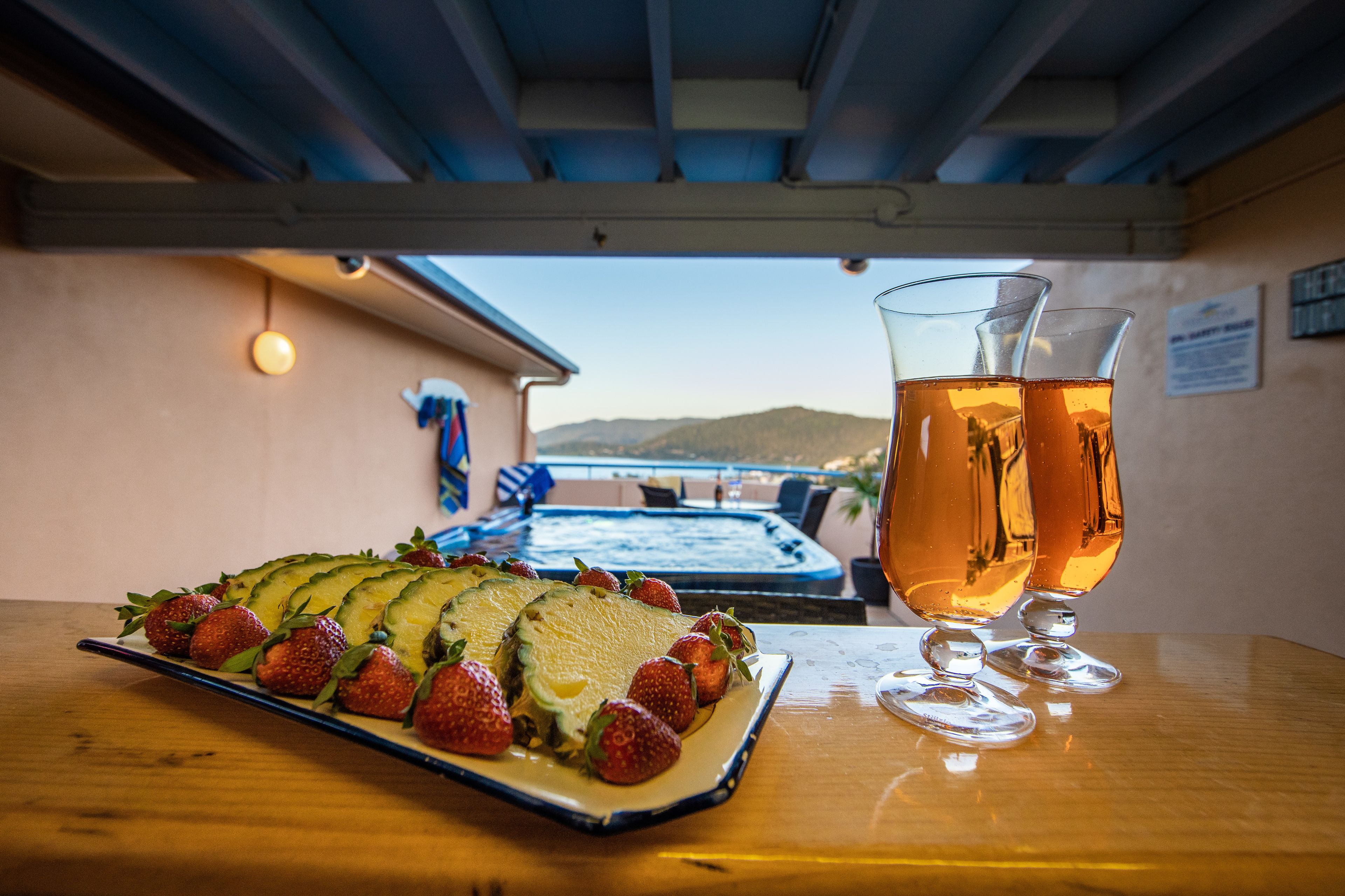 A table with a sliced avocado and strawberries, two glasses of a golden beverage, and a view of a pool and mountains in the background.