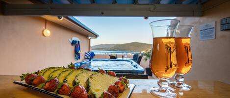 A table with a sliced avocado and strawberries, two glasses of a golden beverage, and a view of a pool and mountains in the background.