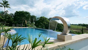 A swimming pool with a modern water feature, surrounded by lush greenery and a clear sky.