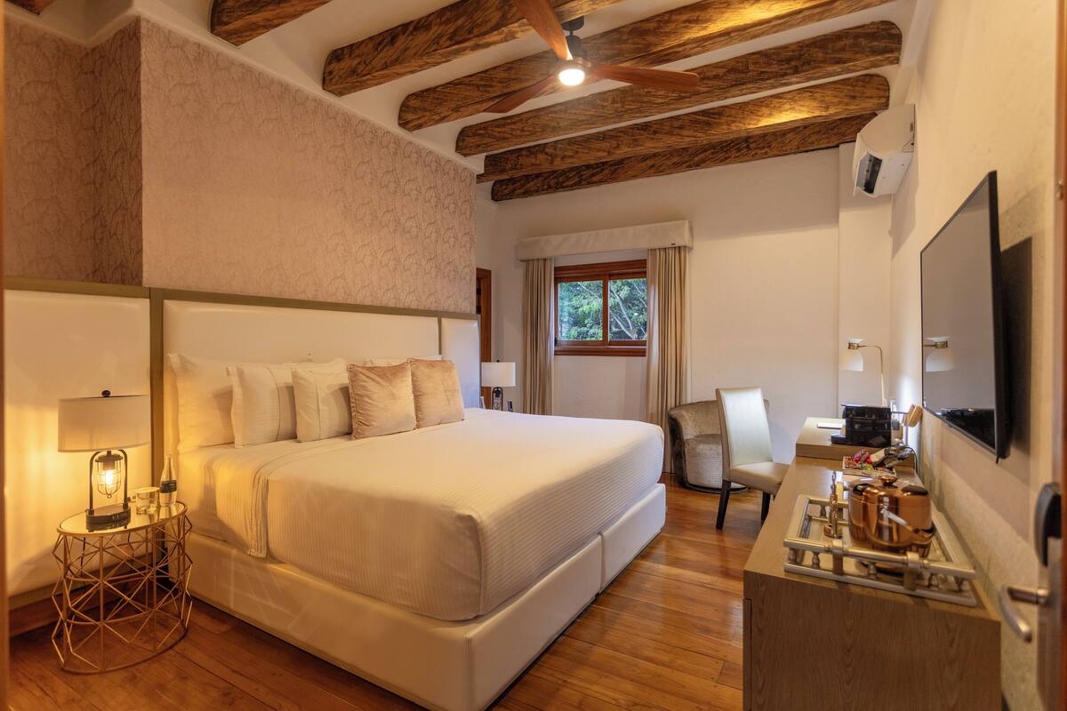 Un dormitorio con una cama grande, un escritorio con cafetera, una silla, un ventanal con cortinas y vigas de madera en el techo.