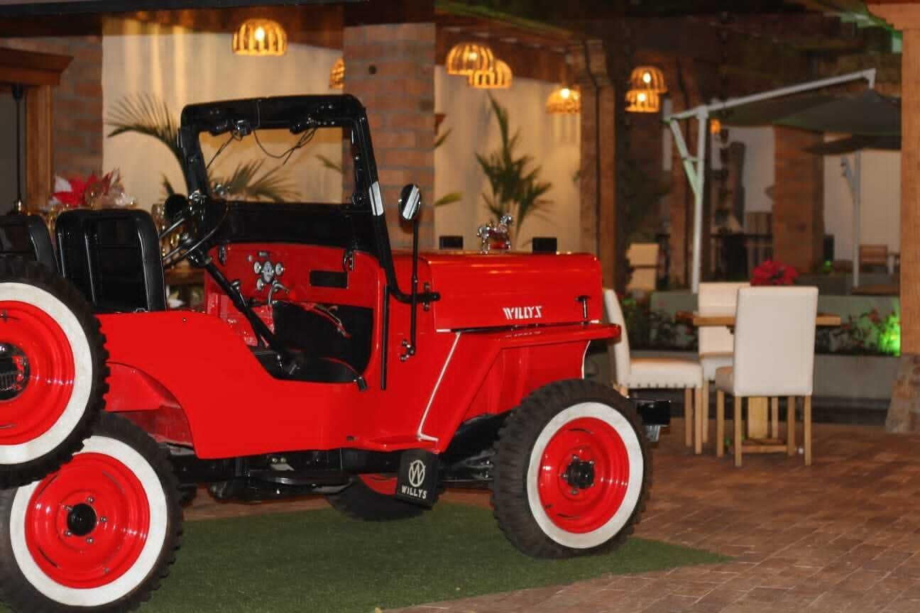 A red Willys off-road vehicle parked in an outdoor area with a brick wall and indoor seating in the background.
