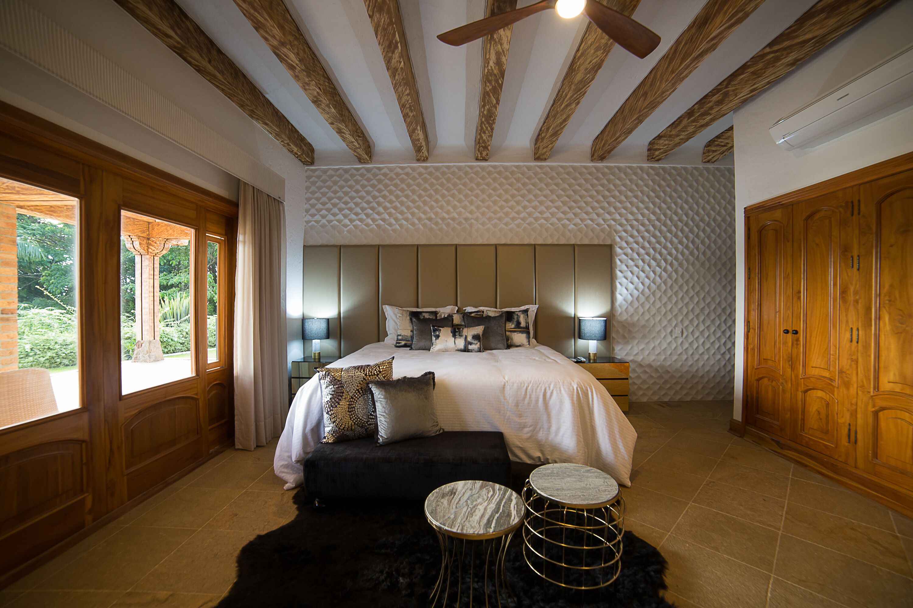Quarto com cama grande, abajures, teto de madeira e vista para o verde lá fora.