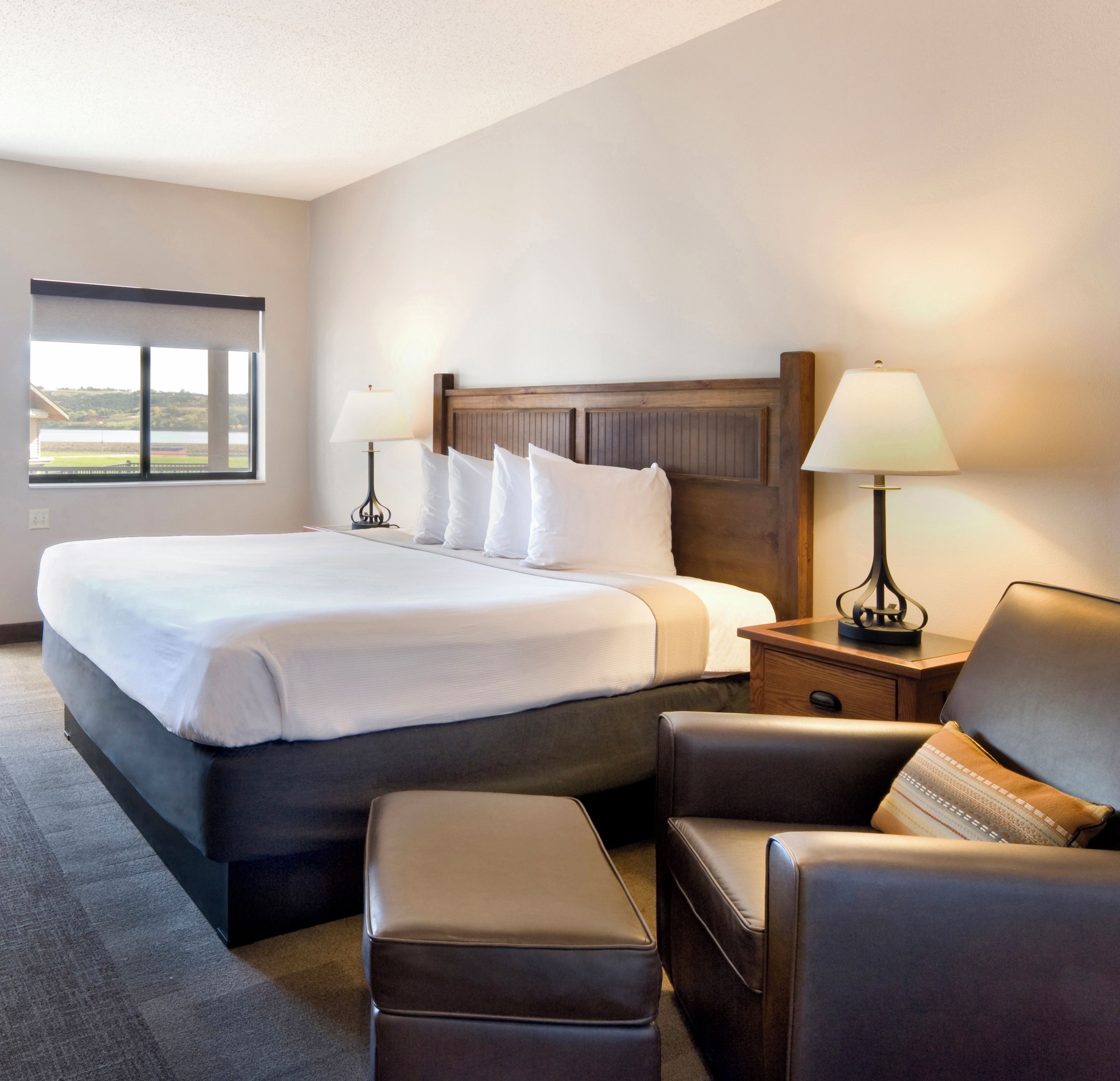 A hotel room with a large bed, two bedside tables with lamps, a chair, and a view of a body of water through a window.