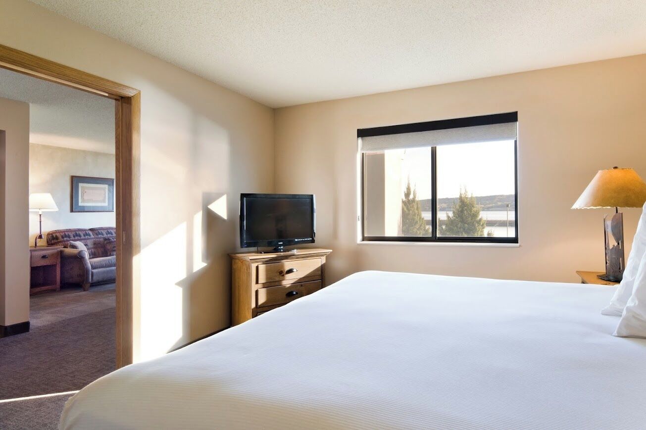 A hotel room with a large bed, a television on a wooden dresser, a window with a view, and a lamp on the side table.