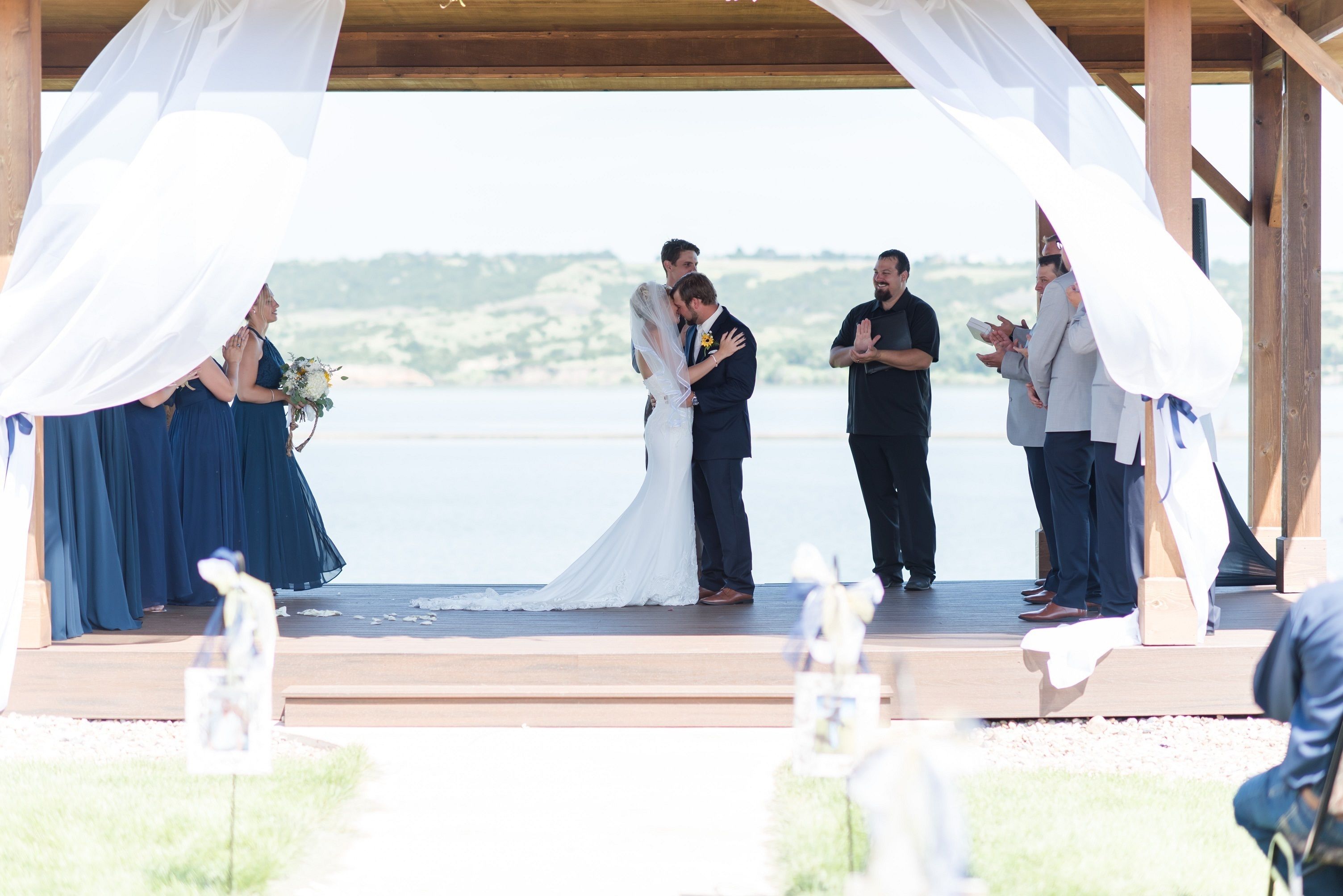 A wedding ceremony on a wooden platform with a backdrop of a body of water and hills.