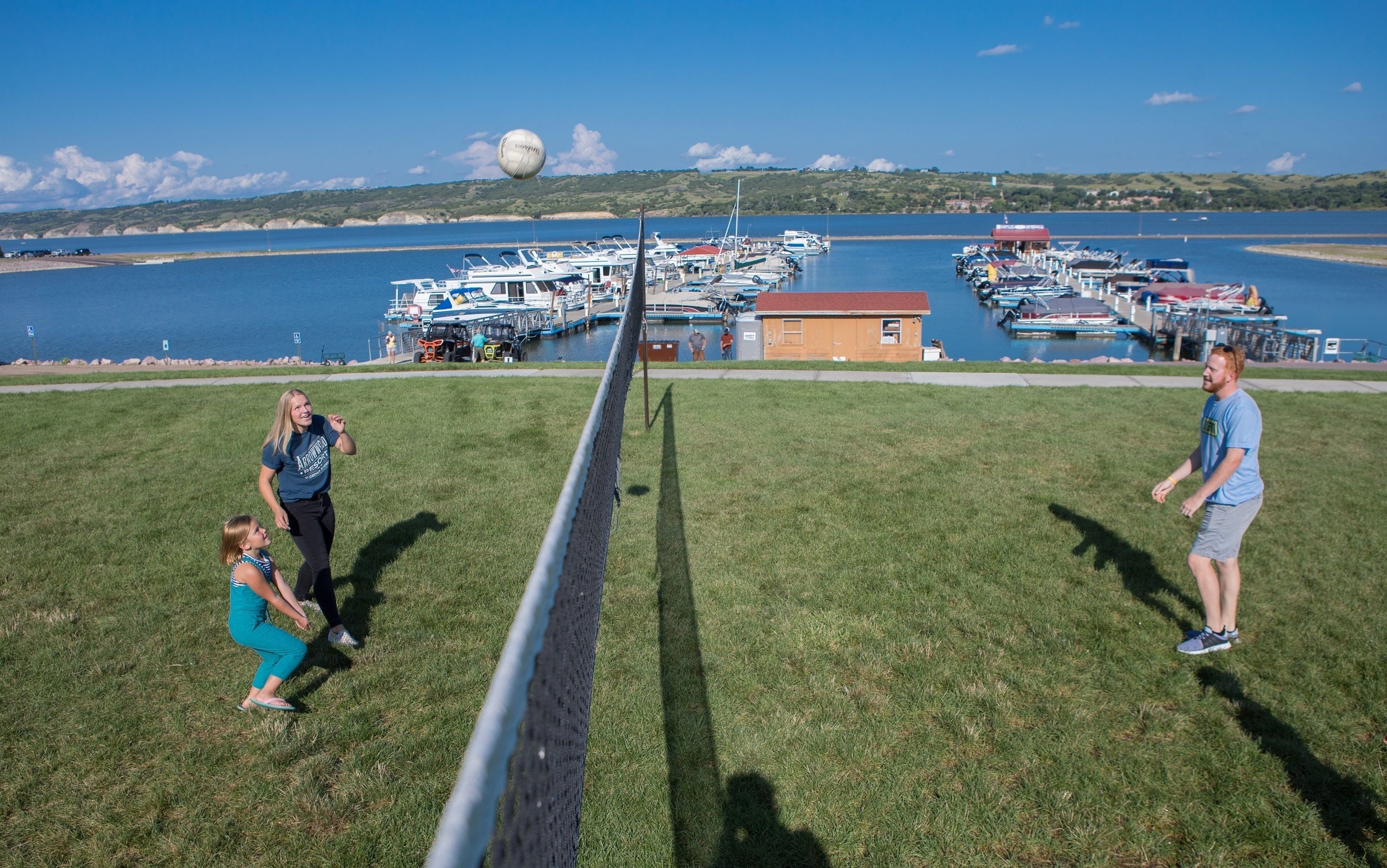 A grassy field with a volleyball net, a small building, and a marina with boats in the background.
