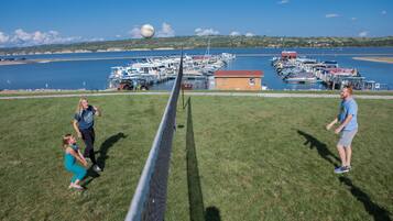 A grassy field with a volleyball net, a small building, and a marina with boats in the background.