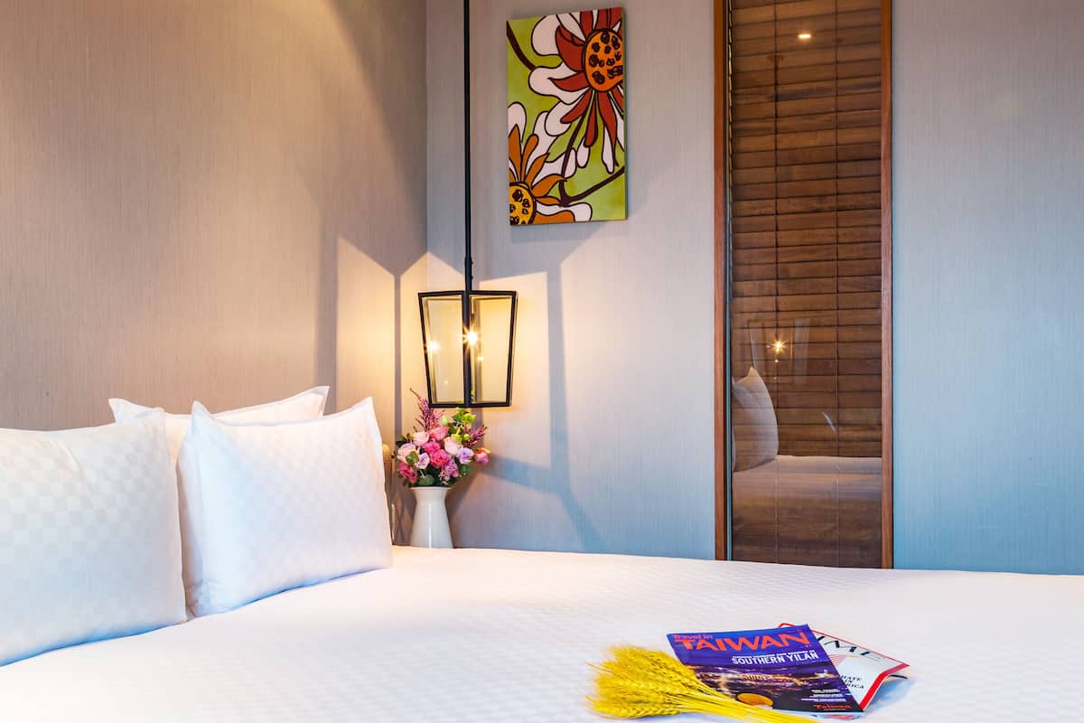 A hotel room with a bed, pillows, a vase of flowers, a framed picture on the wall, and a magazine on the bed.
