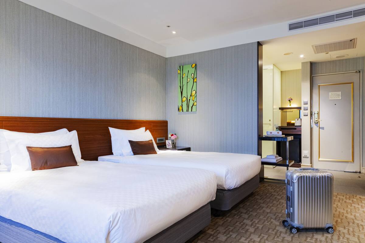 A hotel room with two beds, a wooden headboard, a nightstand, a mirror, a suitcase, and a door to a bathroom.