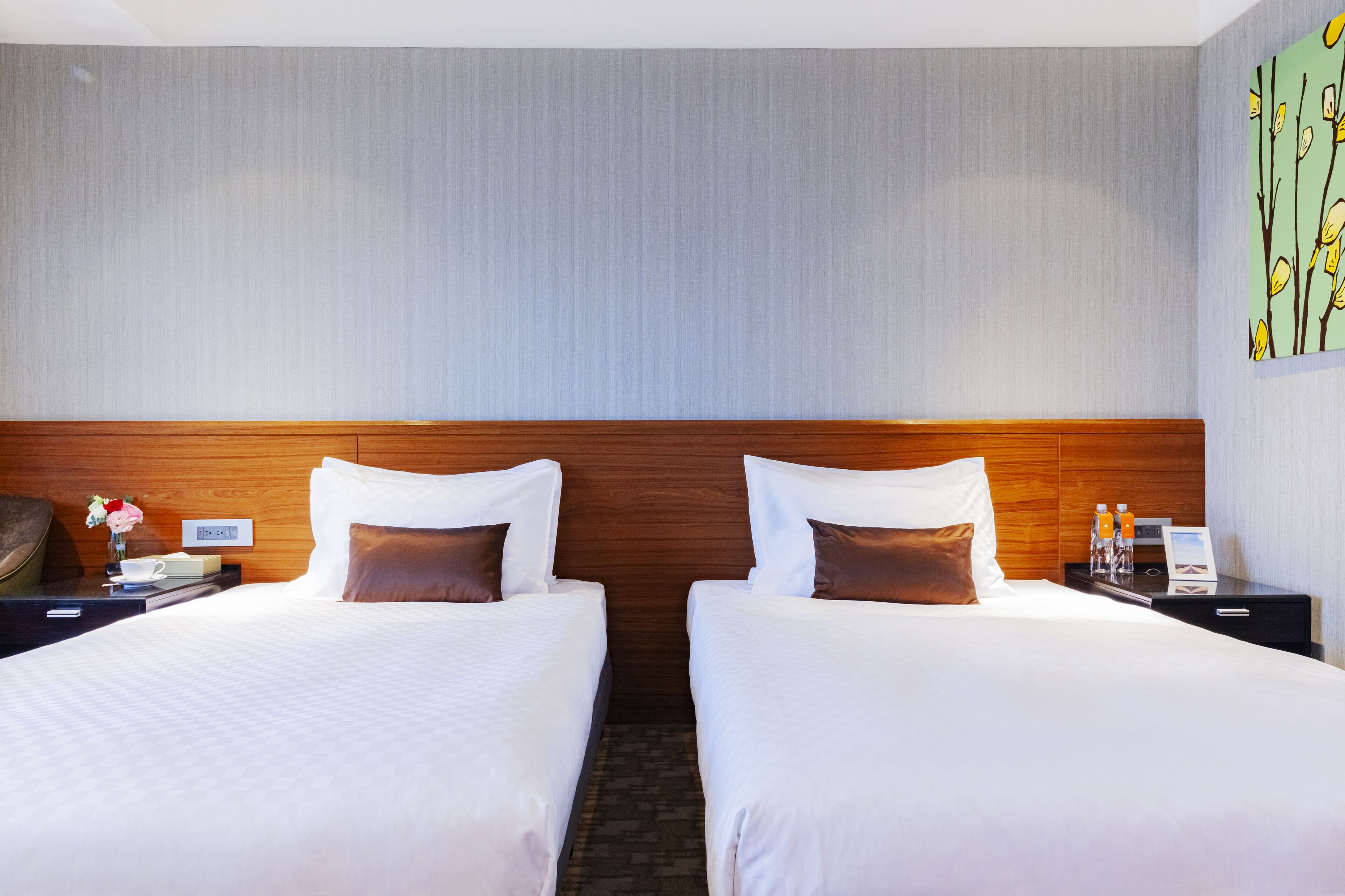 A hotel room with two beds, a wooden headboard, a nightstand with a lamp, and a painting on the wall.