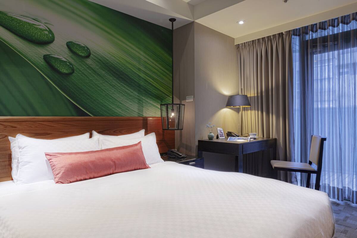 A hotel room with a large bed, a desk with a lamp, a chair, and a view of the outdoors through curtains.