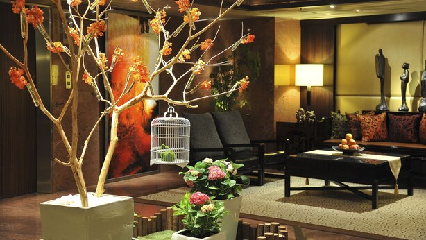 A modern lobby with a large potted tree, a white birdcage, a sofa, and a coffee table with fruit.