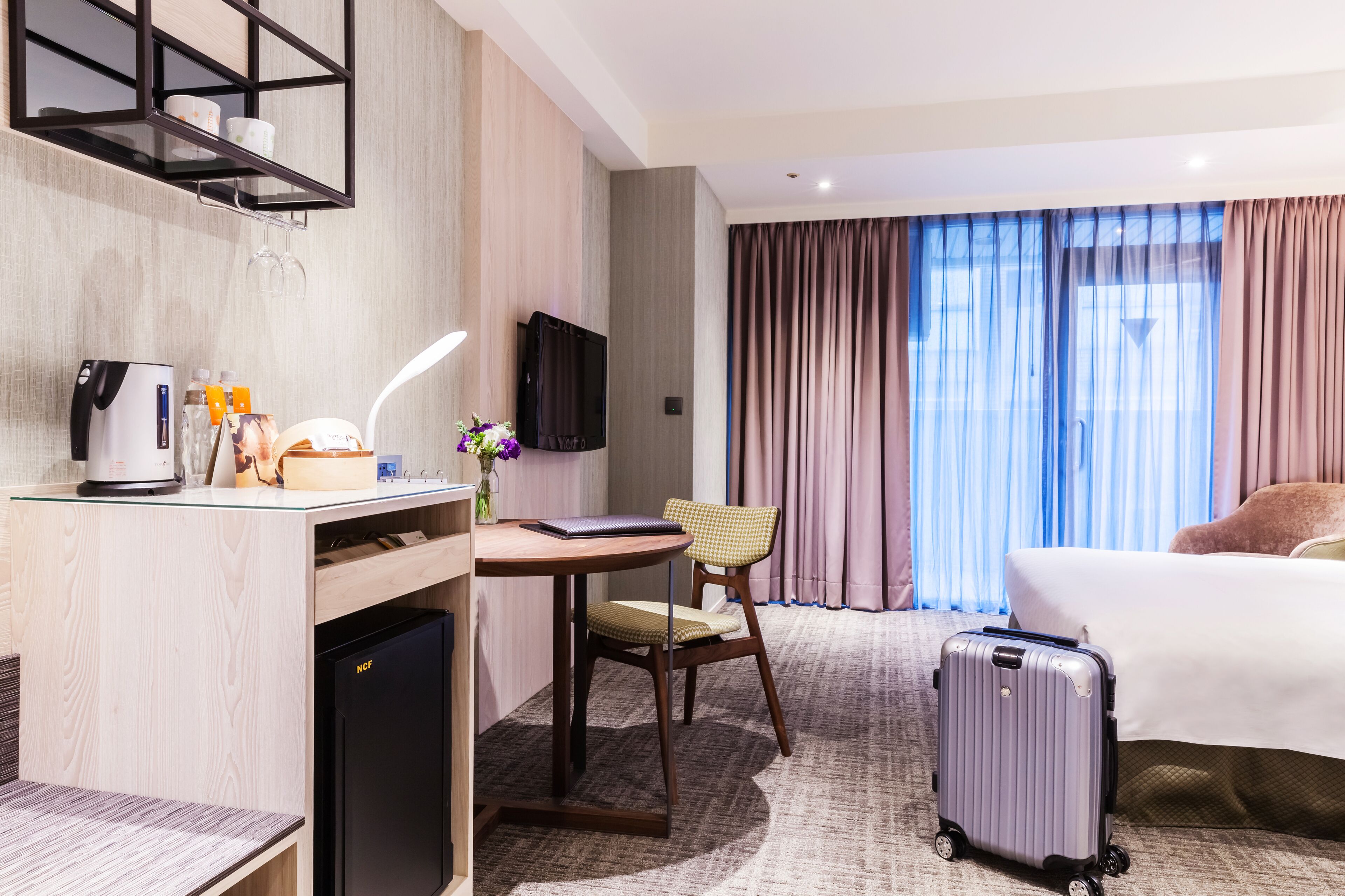 A hotel room with a bed, a desk with a coffee maker, a chair, a suitcase, and a TV on the wall.