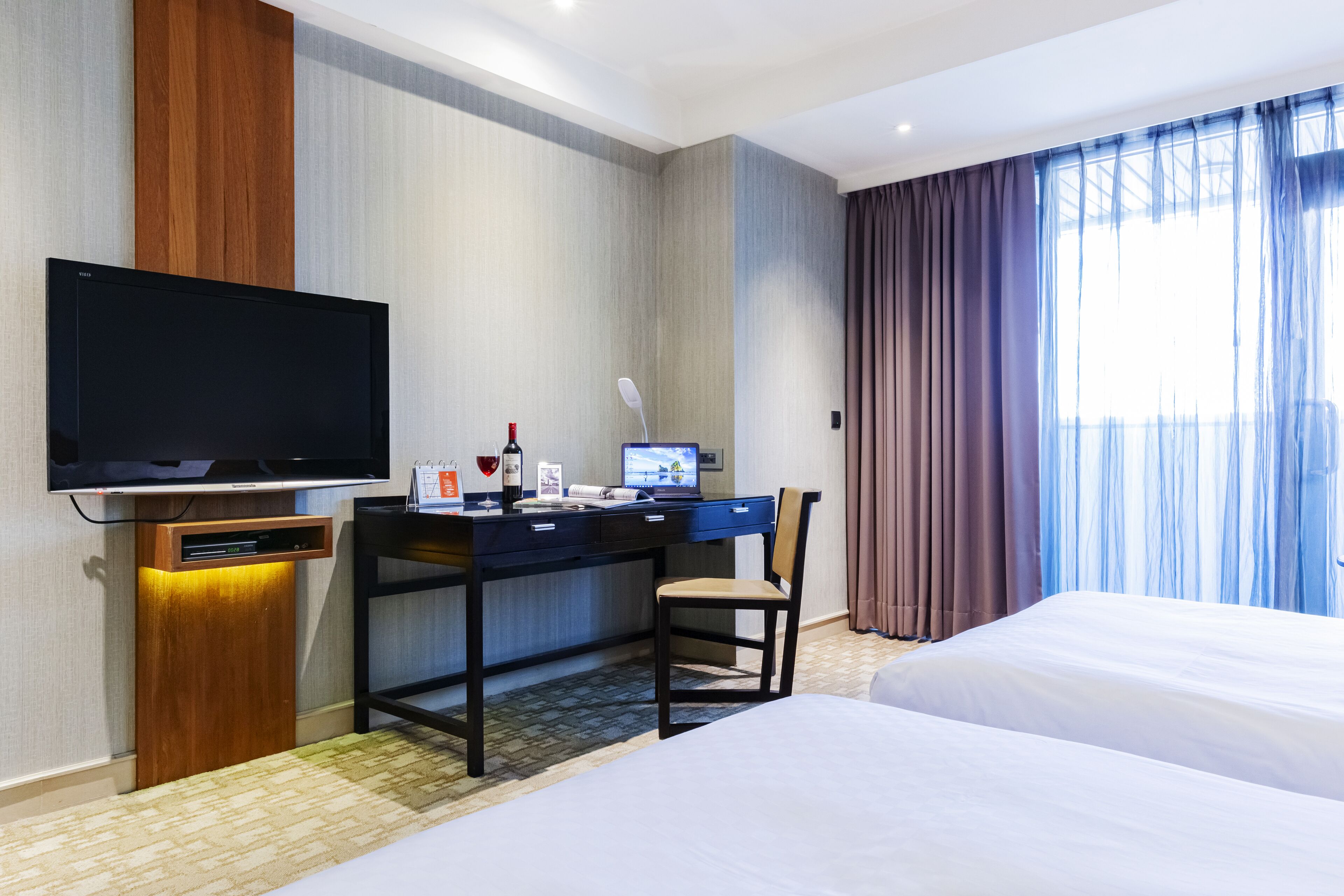 A hotel room with a bed, a desk with a computer, a television, and a window with curtains.
