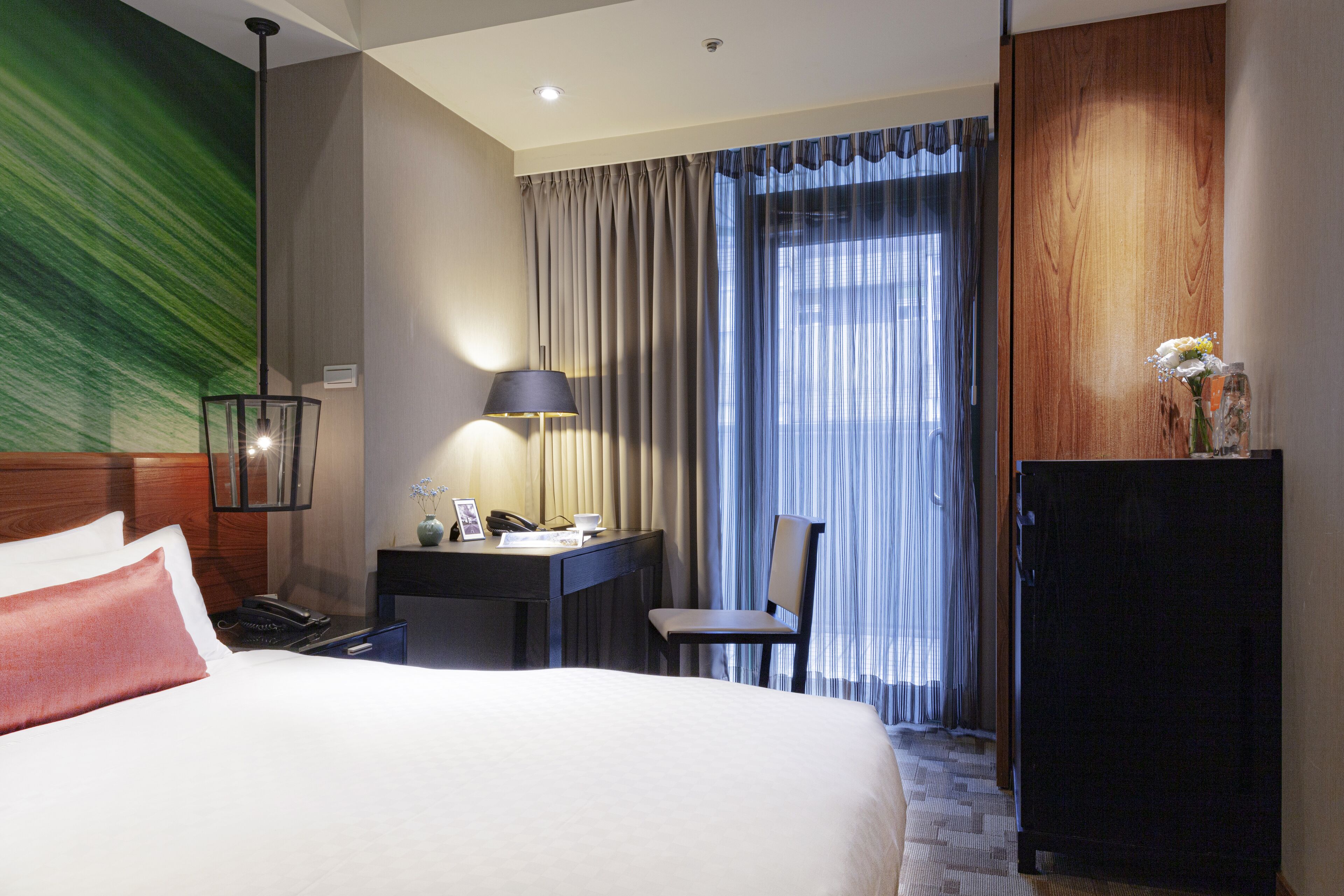 A hotel room with a large bed, a desk with a lamp, a chair, a small table with a vase, and a wardrobe.
