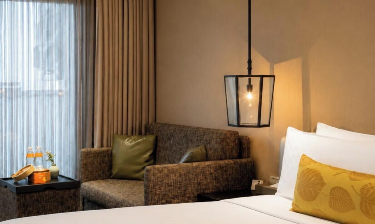 Hotelkamer met een bed, een zithoek met een fauteuil, een nachtkastje met een lamp en een raam met gordijnen.