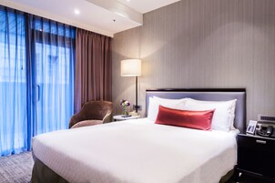 Una habitación de hotel con una cama grande, una silla, una mesita de noche con una lámpara y una mesita auxiliar con un teléfono y un marco de fotos.