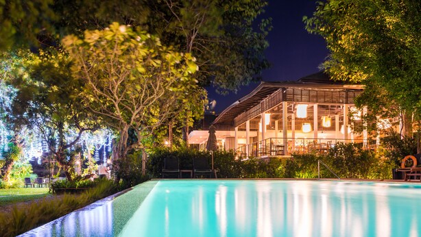A swimming pool with a view of a building with large windows and a covered outdoor area. - The River Resort (Champasak)