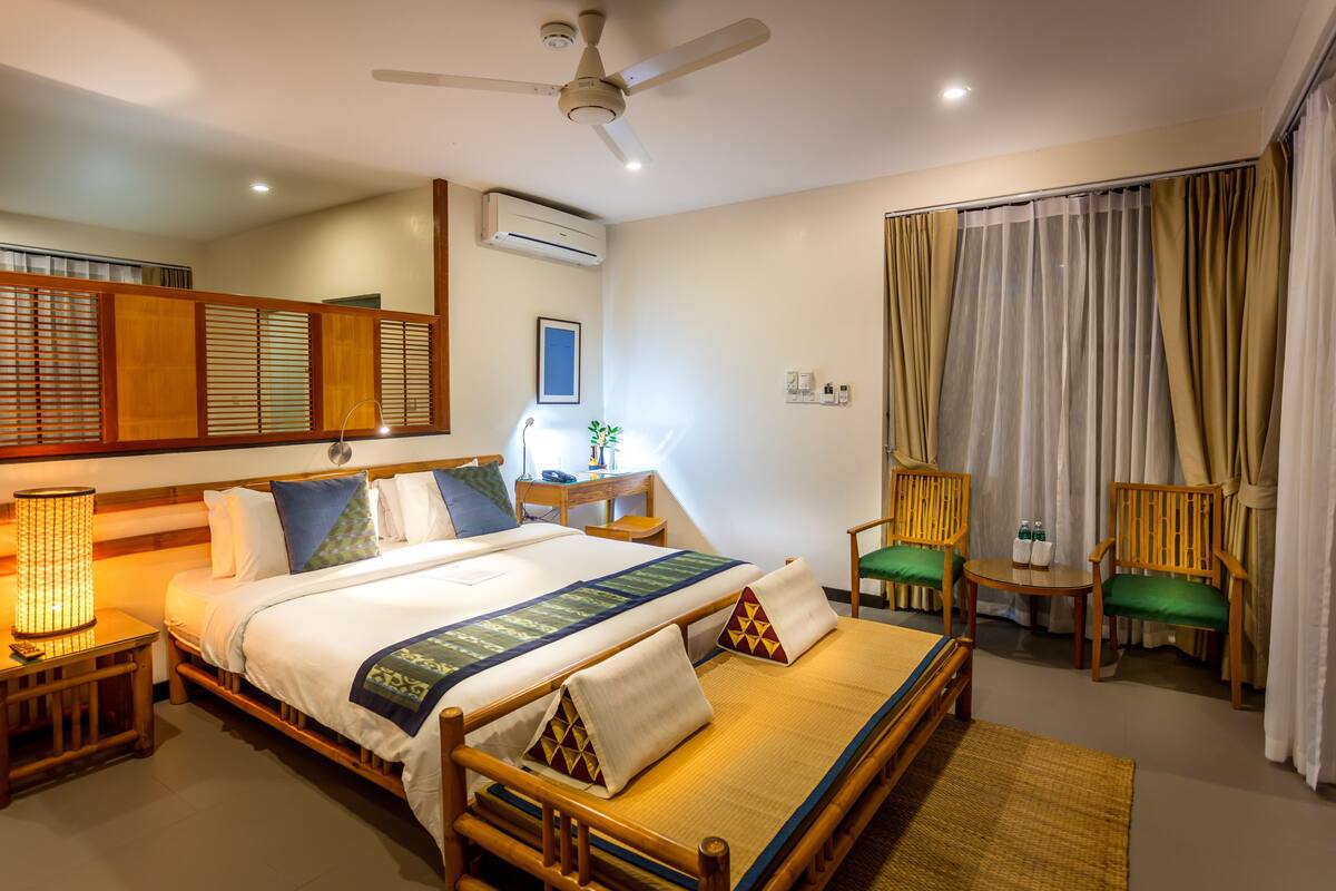 A hotel room with a bed, a nightstand, a lamp, a chair, a table, and a window with curtains.