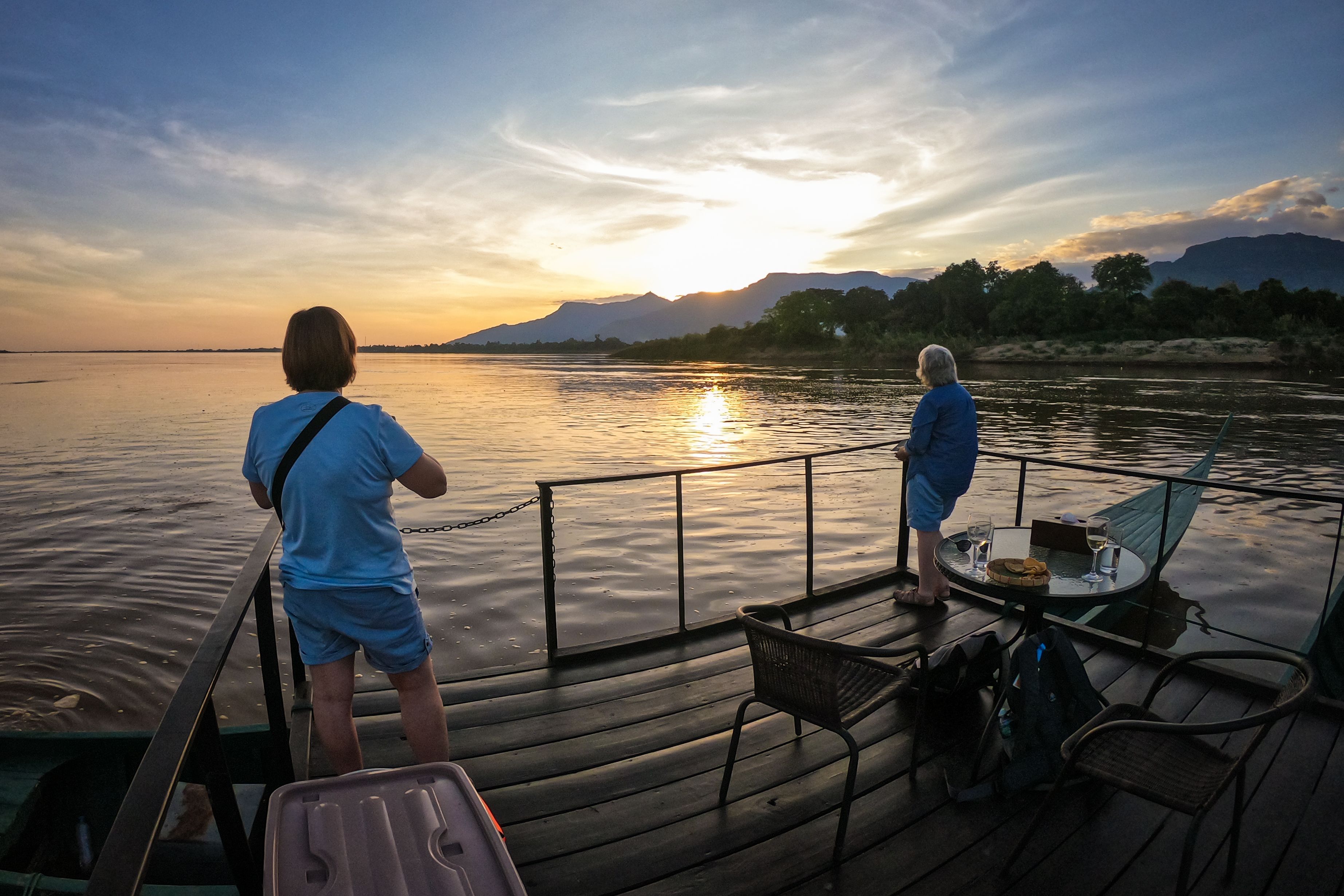 A wooden deck with a table and chairs, overlooking a calm body of water with a sunset in the background.