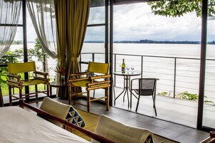 Una habitación con una cama, dos sillas amarillas, una mesita con una botella y vasos, y vista a un cuerpo de agua a través de ventanas amplias.
